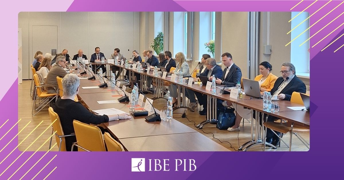Zdjęcie przedstawia uczestników posiedzenia Rady do spraw reformy edukacji zorganizowanego przez Instytut Badań Edukacyjnych. Około 20 osób siedzi przy dużym, prostokątnym stole w sali konferencyjnej. Uczestnicy rozmawiają, notują i korzystają z laptopów. Na stole znajdują się mikrofony, dokumenty, woda oraz tabliczki z imionami i nazwiskami. W tle widoczne są duże okna, rośliny oraz jasne ściany. Zdjęcie oprawione jest w fioletowo-różową grafikę z logo IBE PIB.